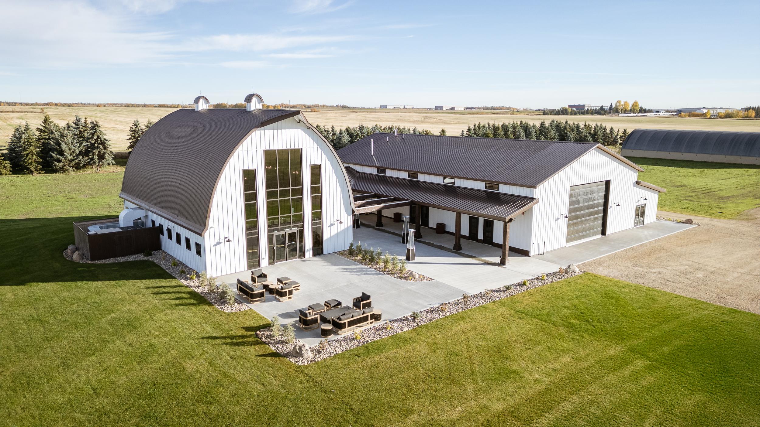 Aerial view of a modern white barn wedding venue with a curved roof, outdoor patio and surrounding green lawn.