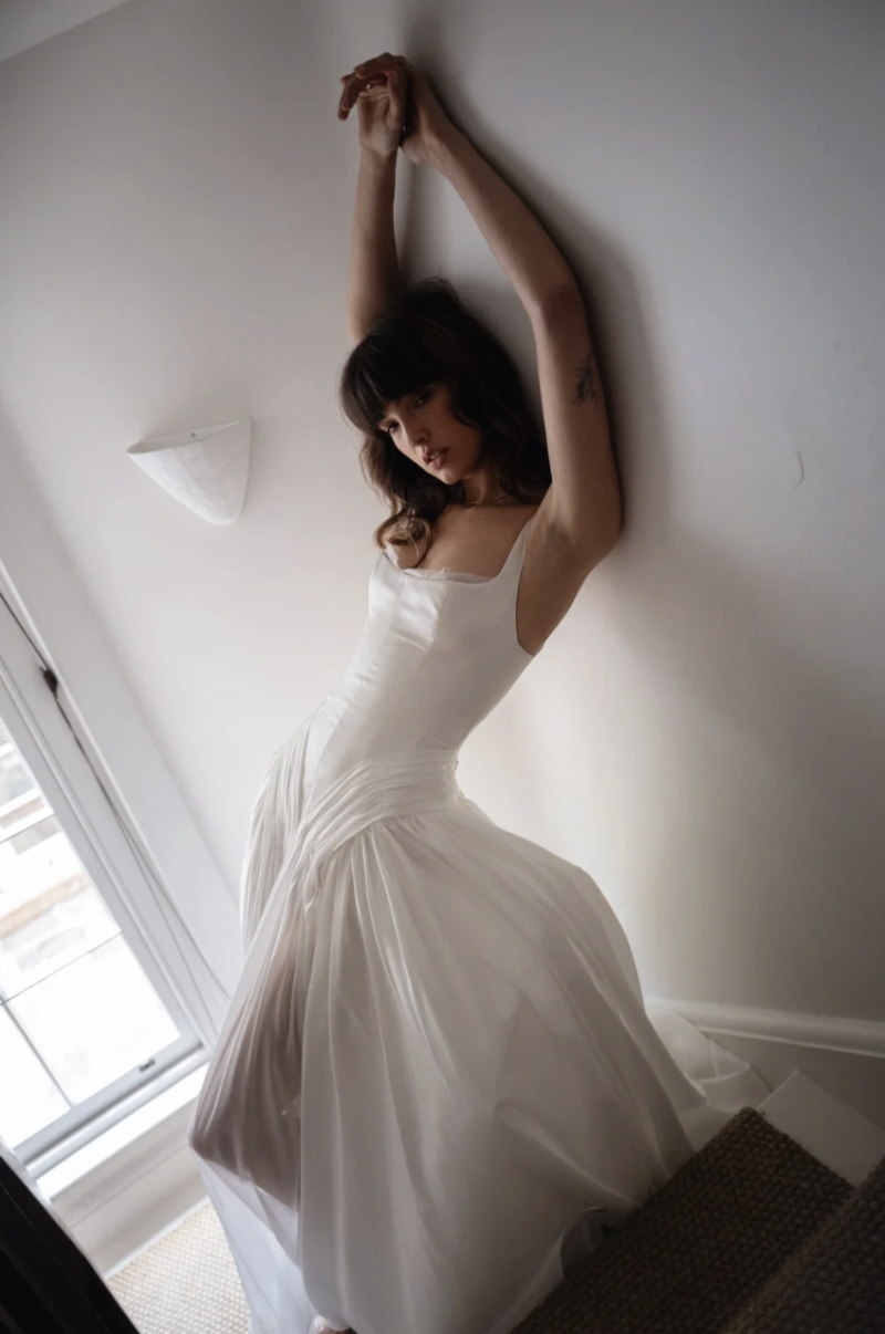 A model wearing the J. Andreatta Citron wedding dress, featuring a softly structured bodice, delicate straps and a full gathered skirt, poses beside a bright window.