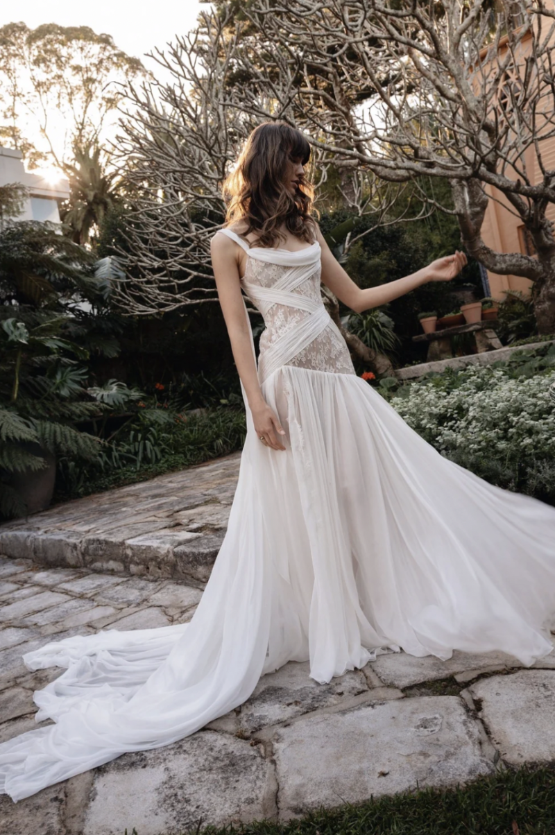 Full-length view of the J. Andreatta Elodie bridal gown with a sheer lace bodice, wrap-style draping, soft skirt and long train photographed on a stone garden path.