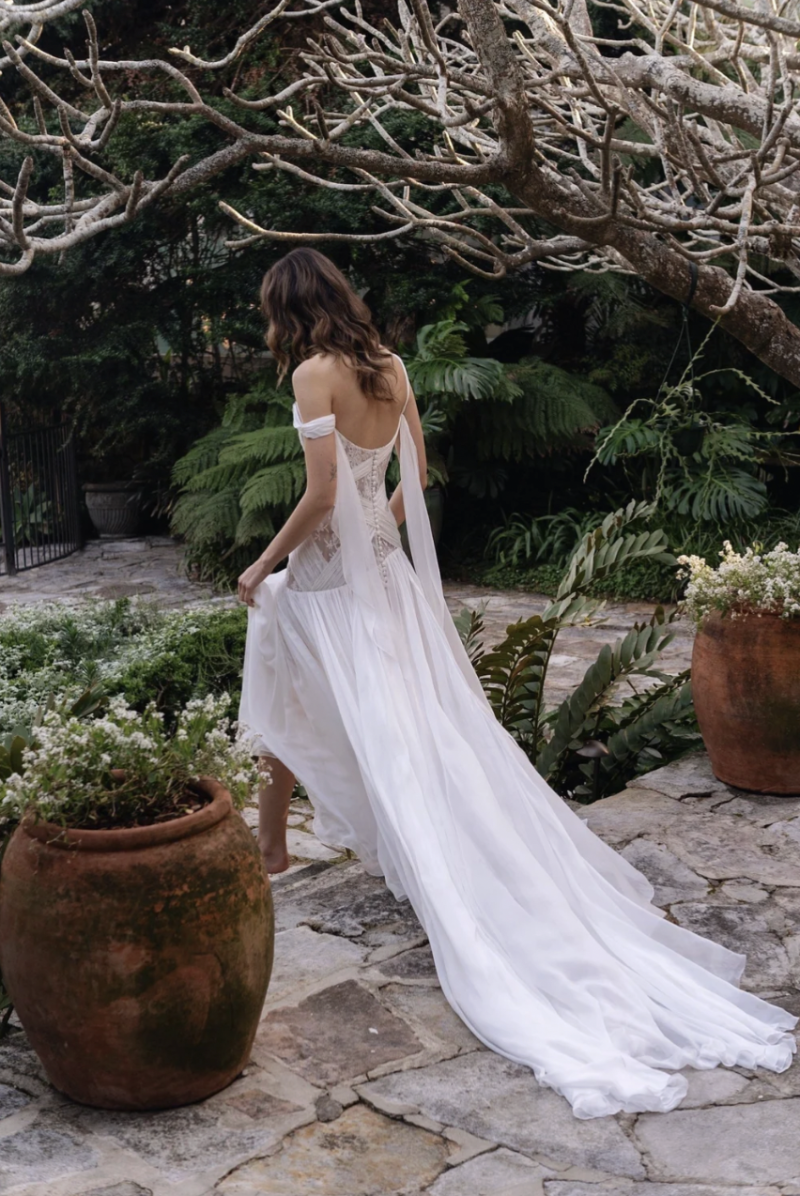 Back view of the J. Andreatta Elodie wedding dress, showing delicate lace details, draped off-the-shoulder fabric and a flowing train in an outdoor garden.