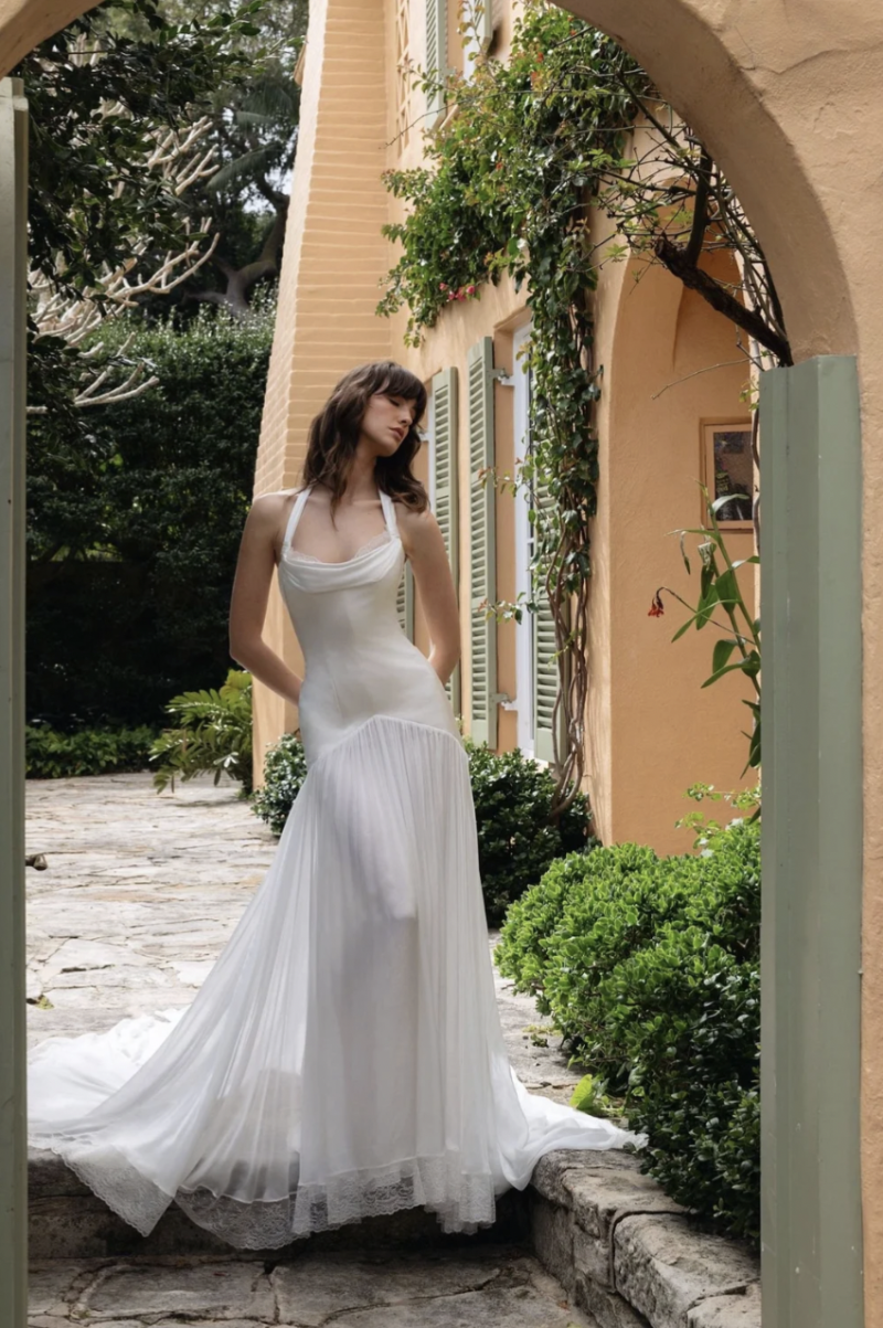 A model wearing the J. Andreatta Indre wedding dress, featuring a fitted bodice, draped neckline, halter-style straps and pleated skirt with lace trim, poses beneath a garden archway.