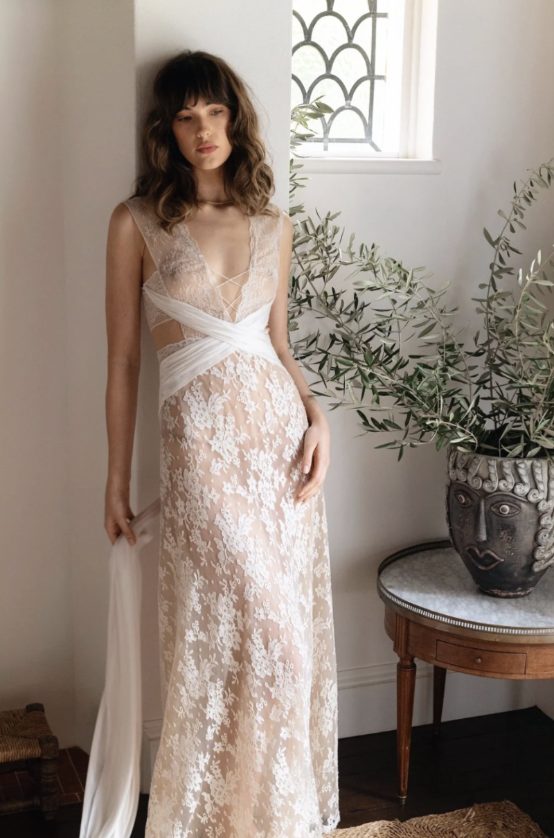 A model wearing the J. Andreatta Lilou wedding dress, featuring sheer floral lace, a plunging neckline, wrap-style draping and soft trailing fabric, poses indoors beside an olive tree.