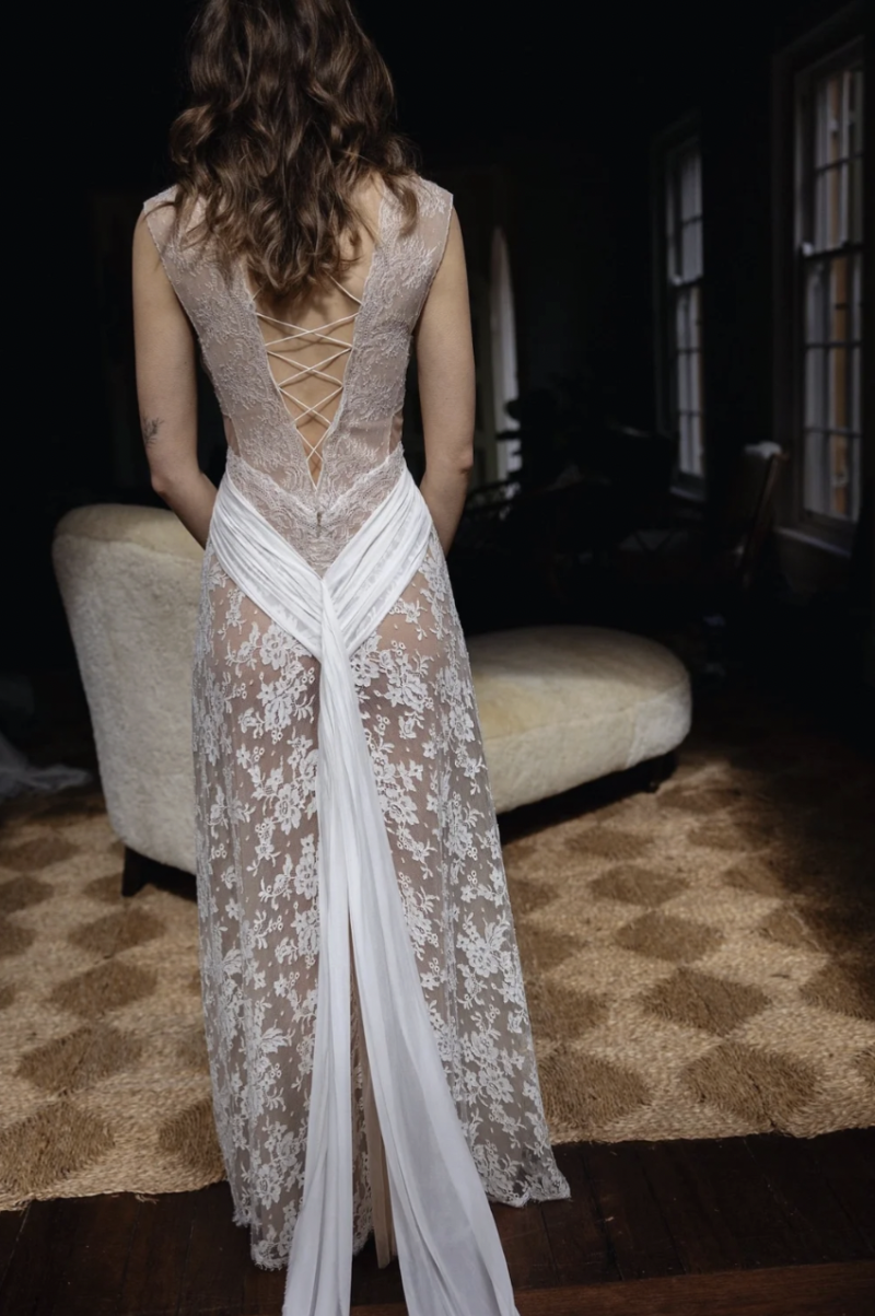 Back view of the J. Andreatta Lilou wedding dress, showing sheer lace, an open lace-up back, wrap-style draping and trailing fabric indoors.