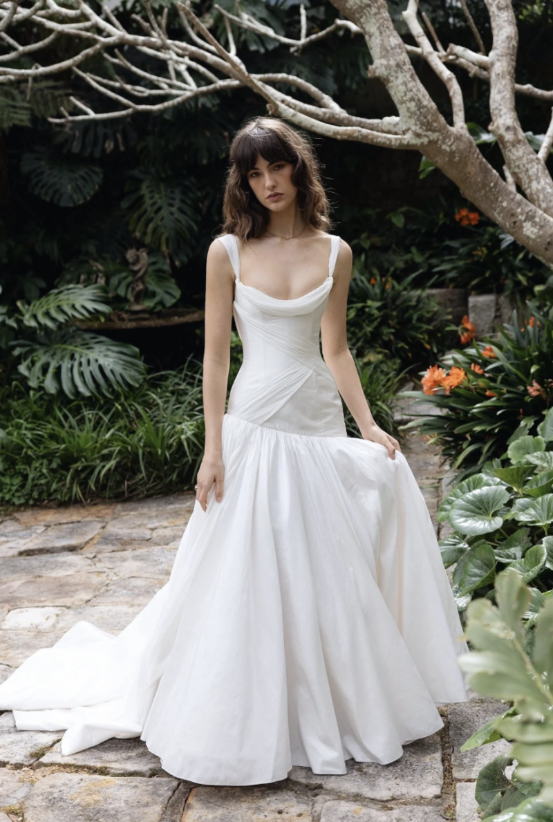 J. Andreatta Nique wedding dress with a draped neckline, fitted bodice and full flowing skirt, photographed in a lush garden setting for a modern bridal editorial.