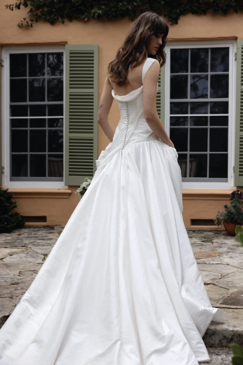 A model wearing the J. Andreatta Nique bridal gown, featuring a low back, draped bodice and voluminous full skirt, poses outside a villa with green shutters.