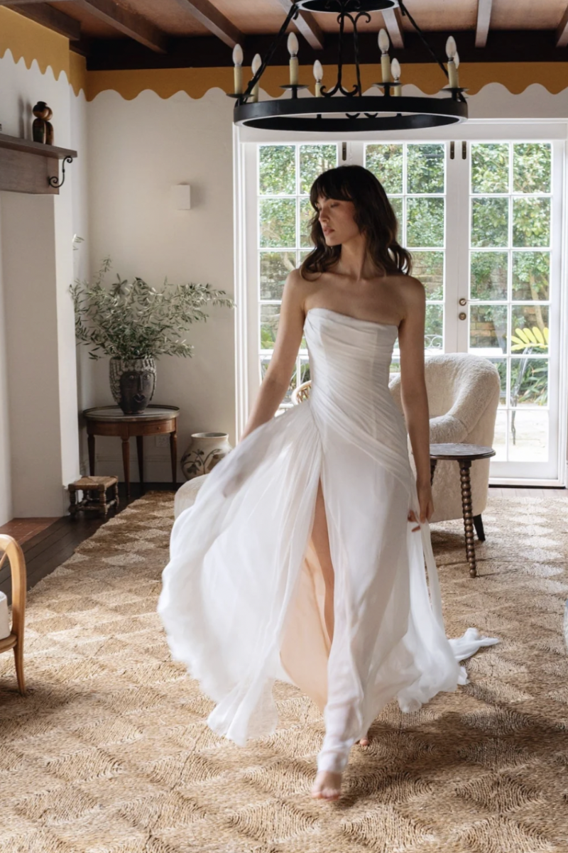 A model wearing the J. Andreatta Ottile wedding dress, featuring a strapless draped bodice, flowing skirt and thigh-high slit, walks through a sunlit interior.