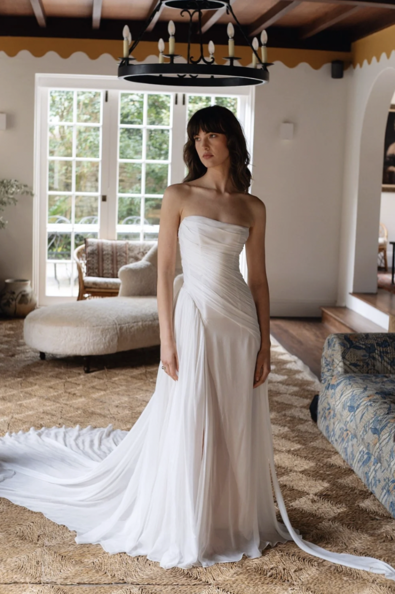Full-length view of the J. Andreatta Ottile bridal gown with a strapless wrap-style bodice, soft pleated skirt and sweeping train photographed indoors.