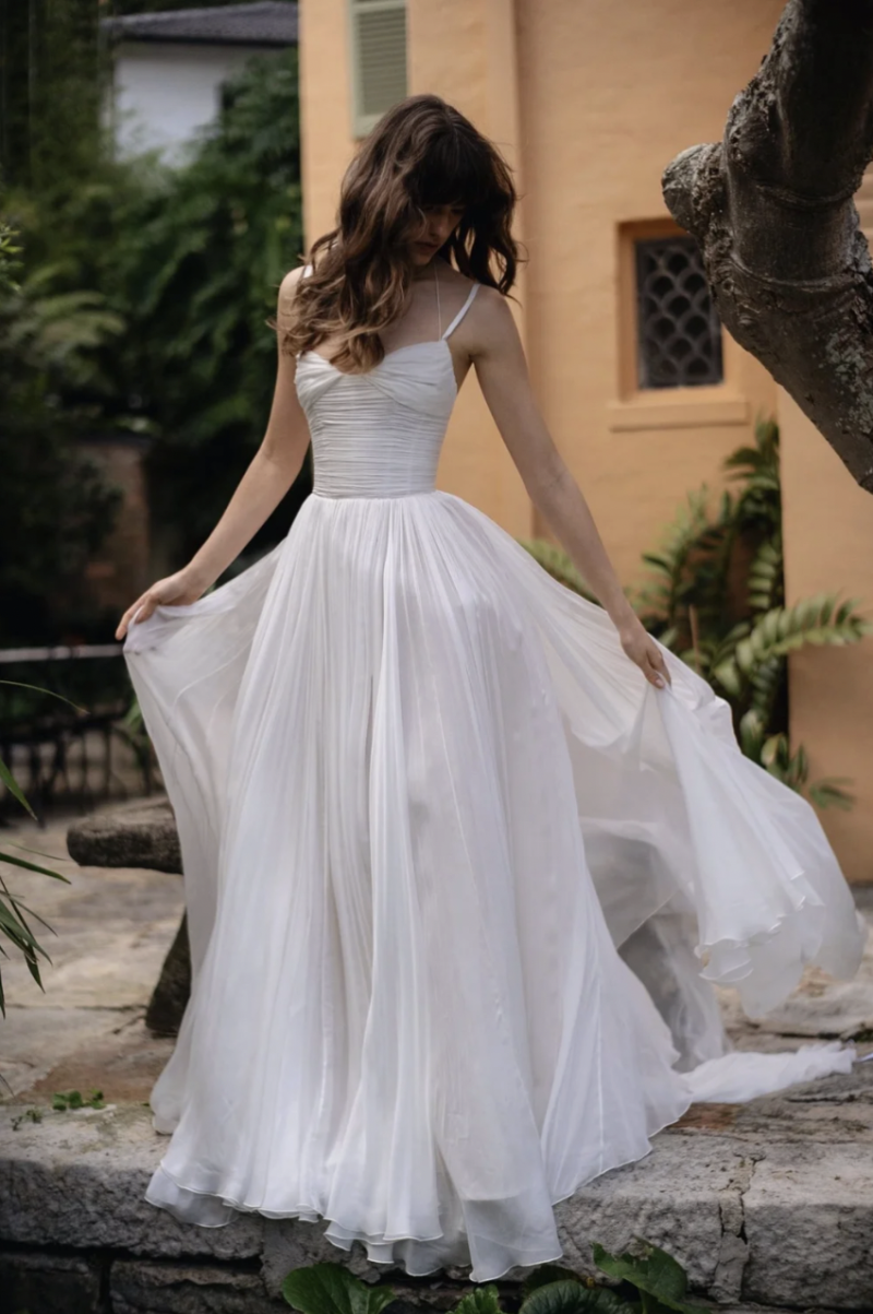 A model wearing the J. Andreatta Silvie wedding dress, featuring a ruched bodice, delicate straps and a full pleated skirt, poses outdoors in a garden setting.