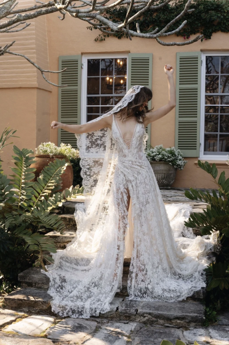 A model wearing the J. Andreatta Soleil bridal look, featuring allover lace, a plunging neckline, hooded veil detail and flowing train, poses on garden steps.