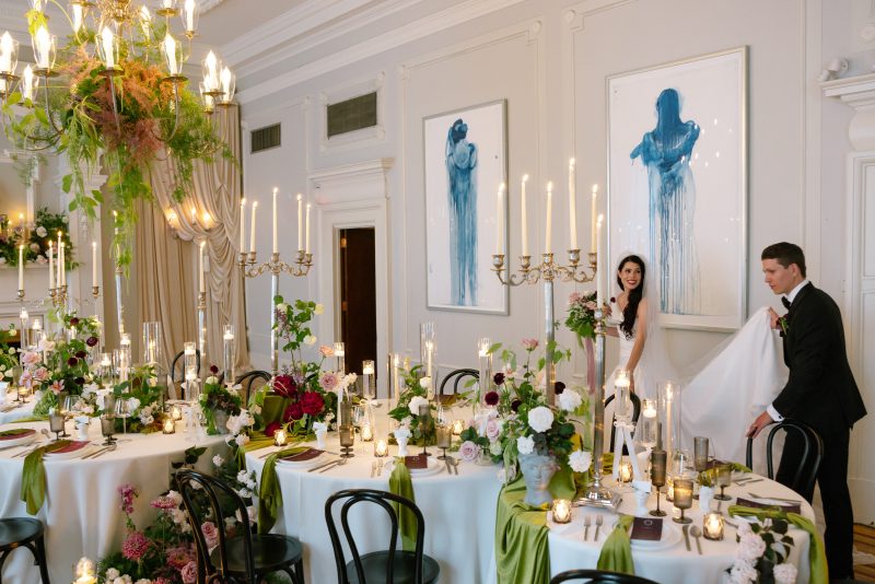 Bride and groom stand beside a candlelit wedding reception at The Vancouver Club, featuring round tables, green velvet napkins, black chairs, tall candelabras and moody romantic florals.