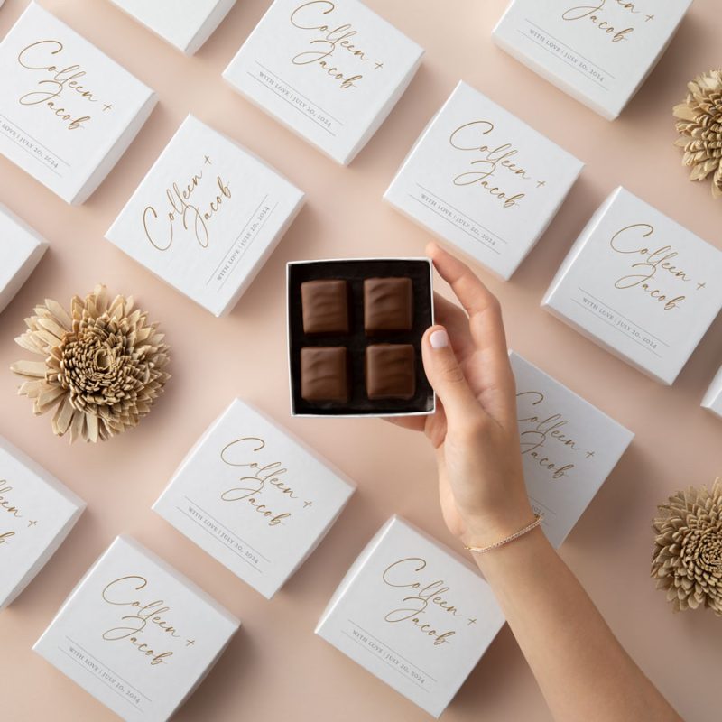 Custom white wedding favor boxes with gold lettering arranged on a blush surface, with one open box revealing four chocolate toffee pieces.