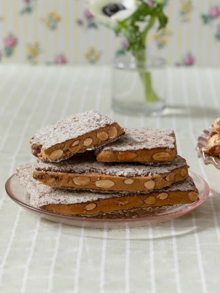 Stacked almond toffee slices dusted with crushed nuts on a pink glass plate with flowers in the background.
