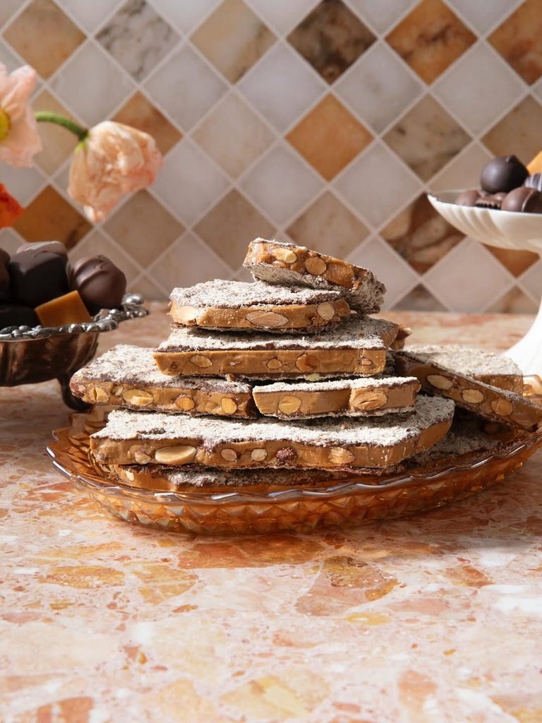 Almond toffee slices stacked on a glass platter against a warm peach marble and tiled backdrop.