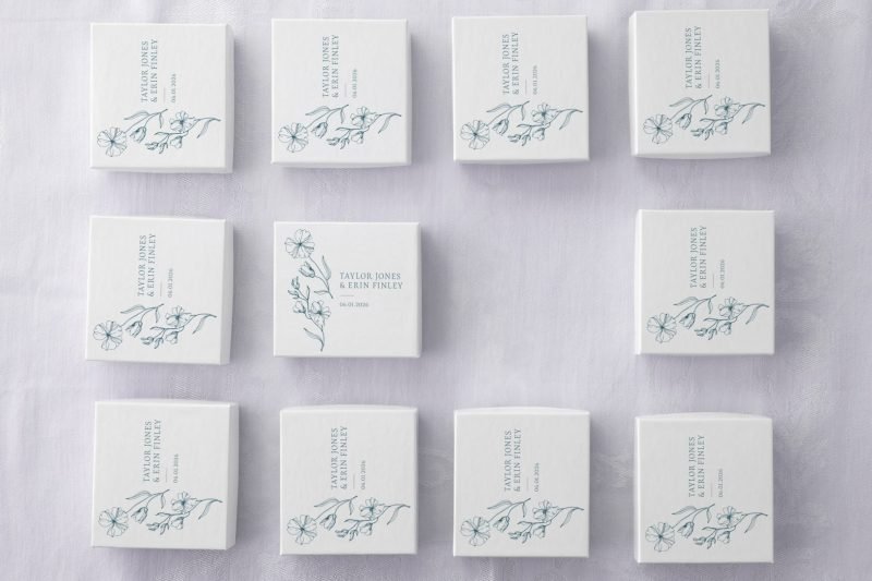 White custom wedding favor boxes with blue floral illustrations and names arranged on a white linen surface.