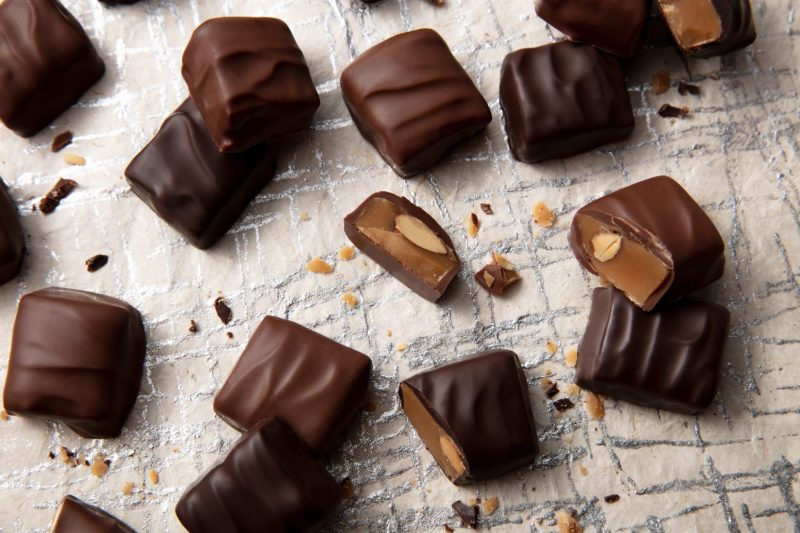 Chocolate-covered almond toffee pieces and sliced pieces with visible almond centers scattered across a textured silver surface.